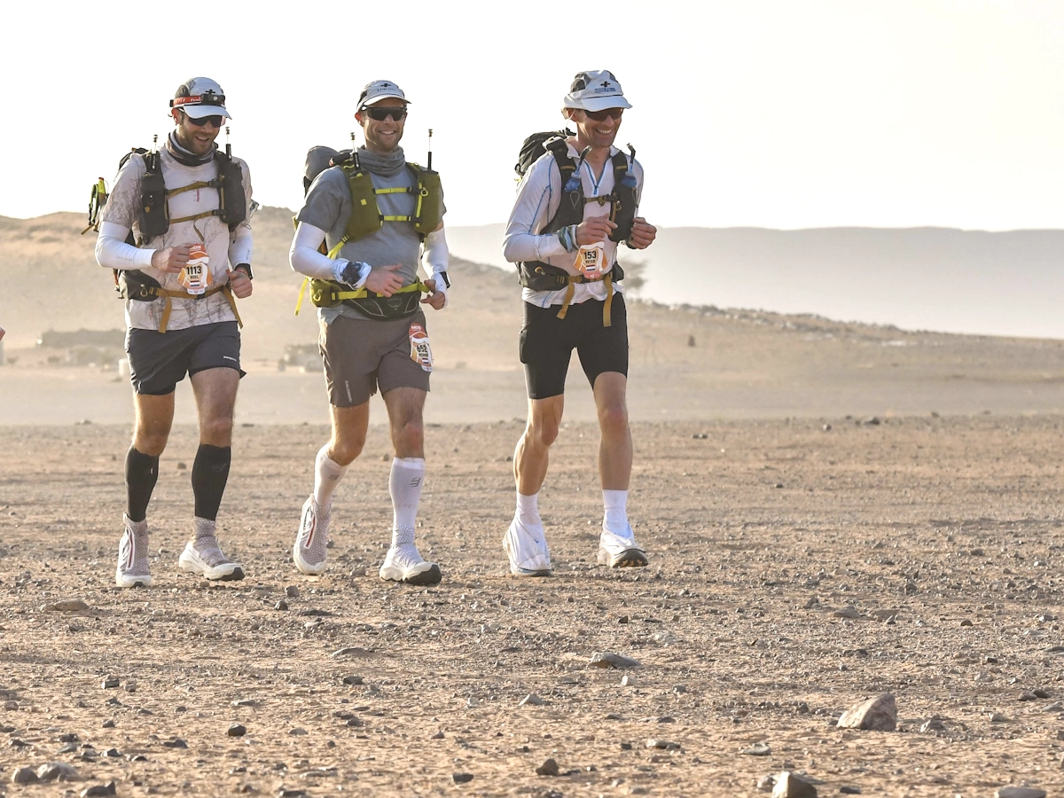 Drie lopers rennen lachend door de woestijn tijdens de Marathon des Sables